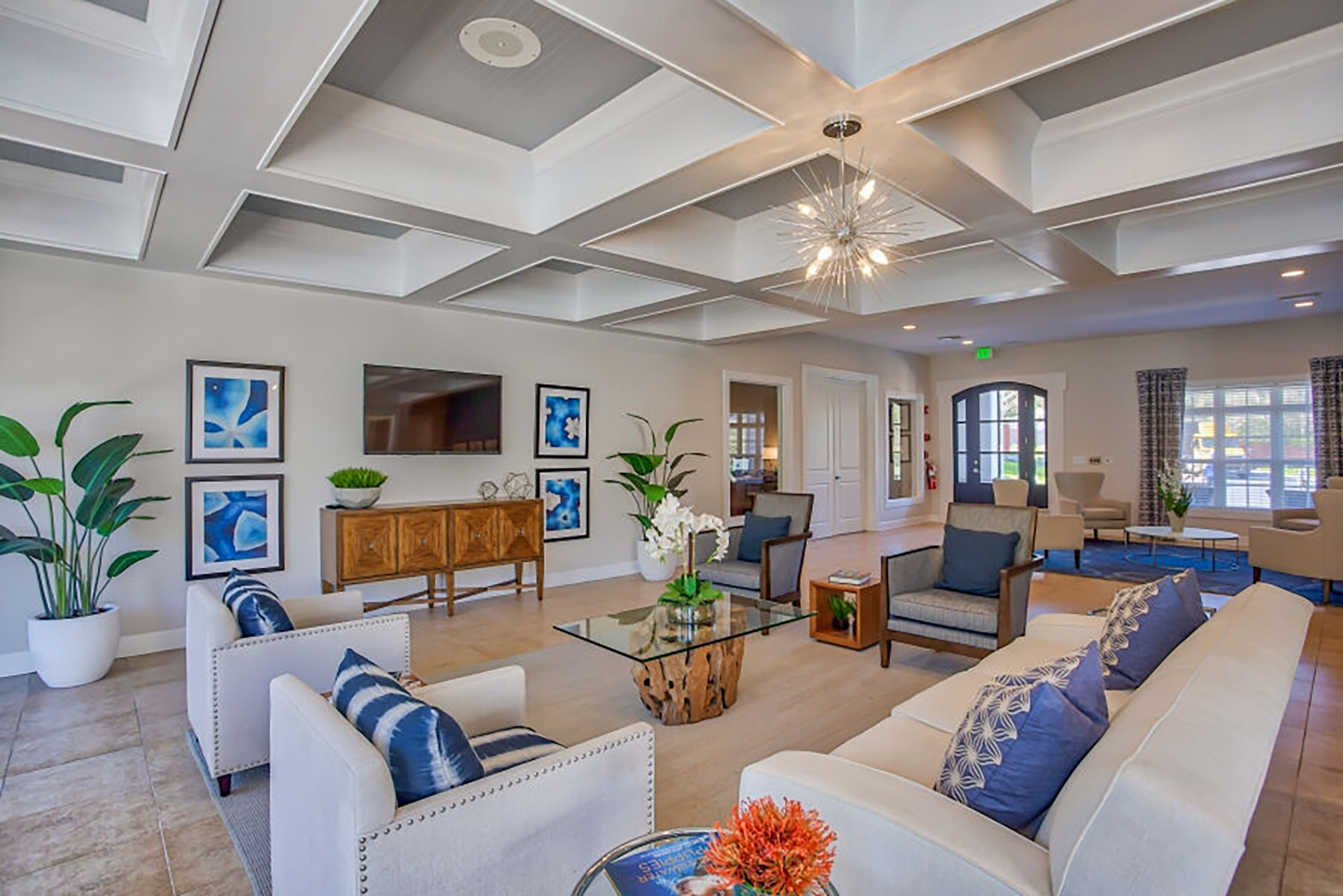 A modern living room with coffered ceiling in a multi-family real estate investment property. Cream-colored sofas, a glass coffee table, and a large TV. Blue accents in pillows and artwork, along with potted plants and a geometric chandelier, enhance the decor.