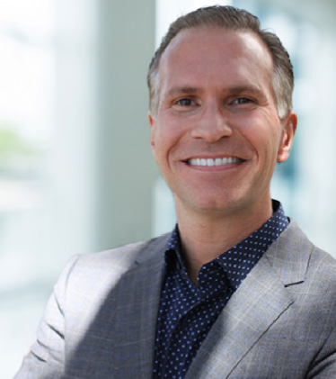 A man in a grey suit jacket and dark blue dotted shirt smiles while standing indoors with a blurred background, representing the professionalism of PEM Real Estate Group Leadership.