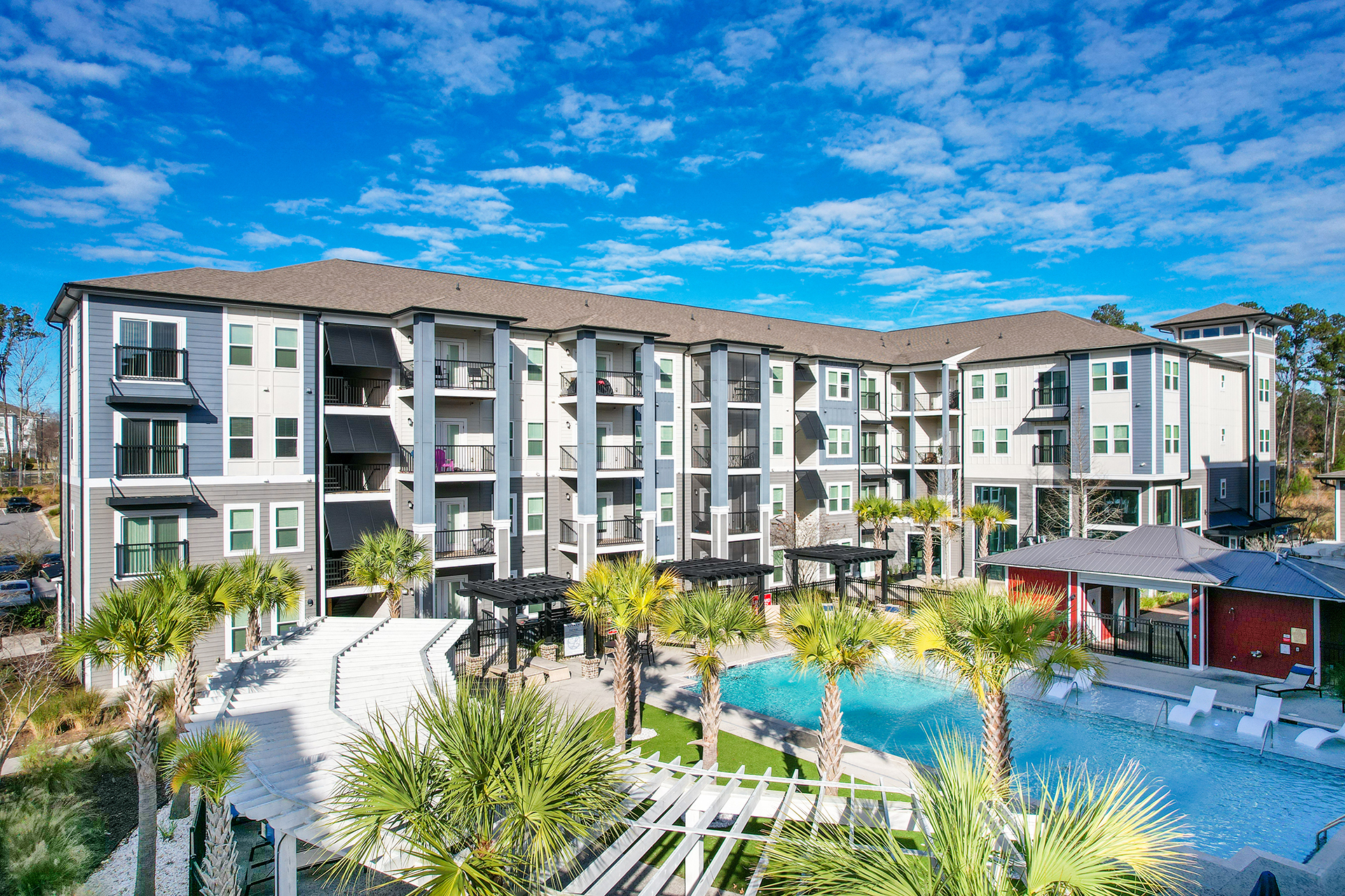 A four-story apartment building with balconies overlooks a swimming pool, surrounded by palm trees and lounge areas, under a bright blue sky—a prime example of multi-family real estate investments.