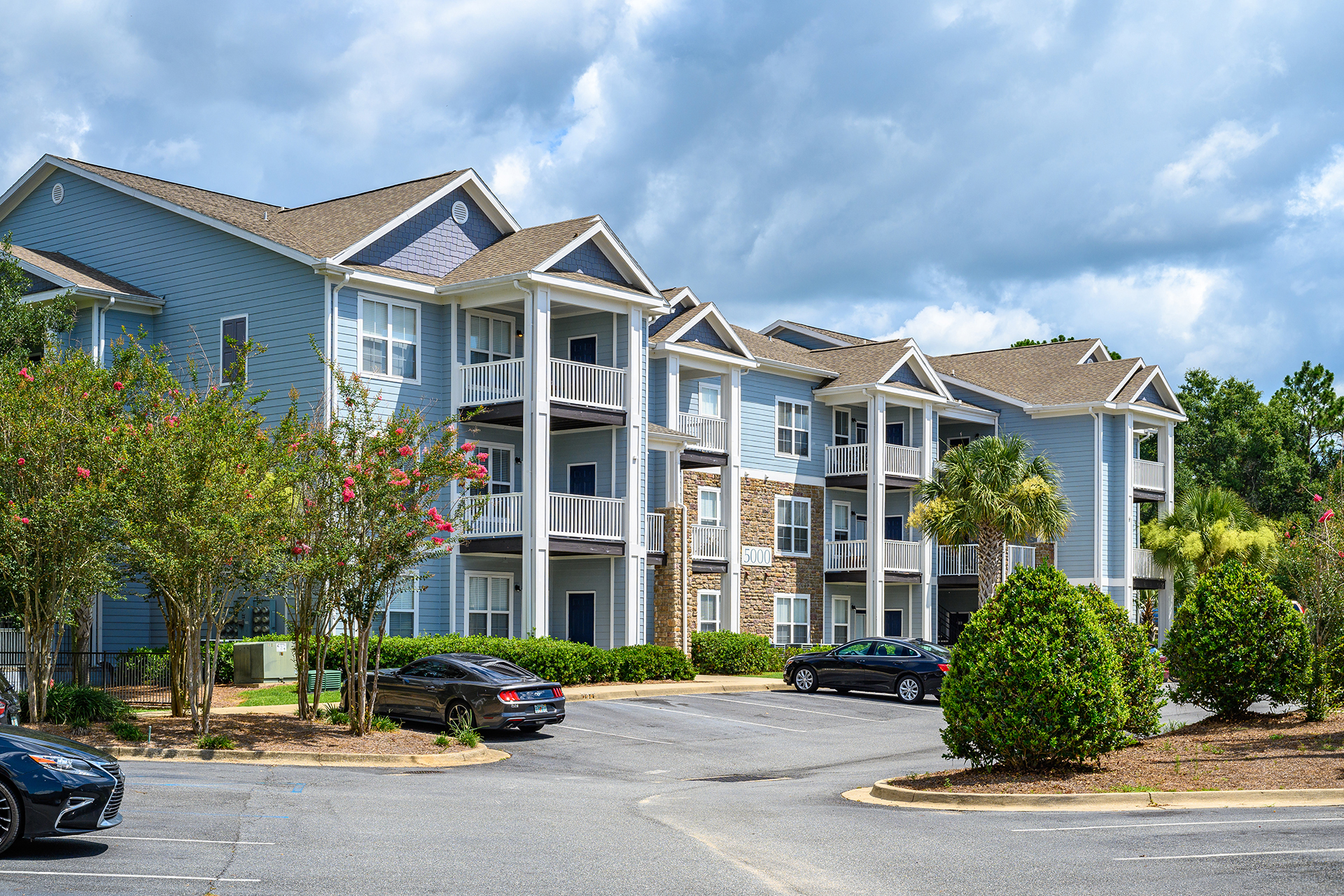 A row of multi-story apartment buildings with balconies, surrounded by trees and shrubbery, epitomizes prime Multi-Family Real Estate Investments. Several cars are parked in the spacious parking lot in the foreground under a partly cloudy sky.